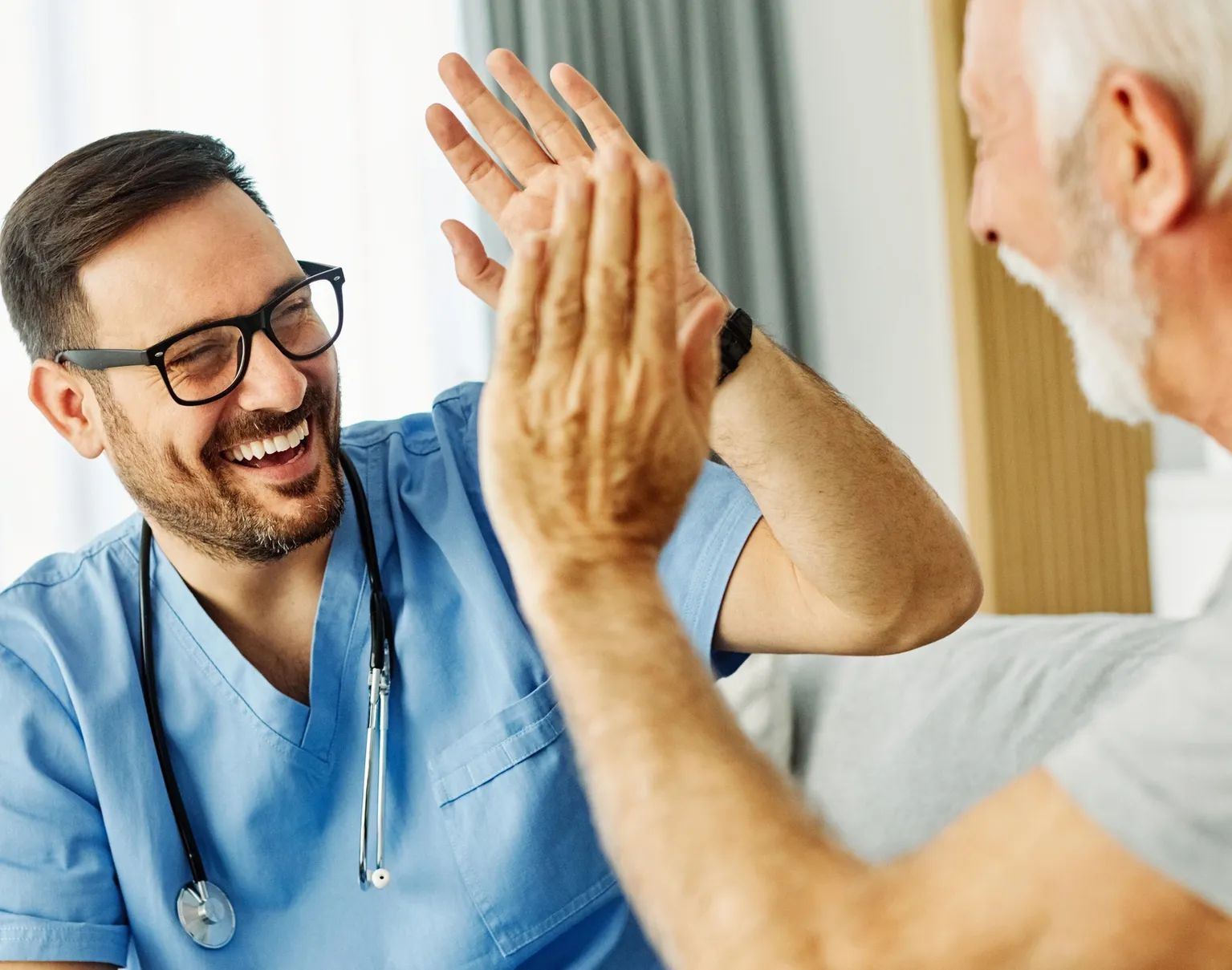 Doctor in blue scrubs giving a high five to an elderly patient. Both are smiling.