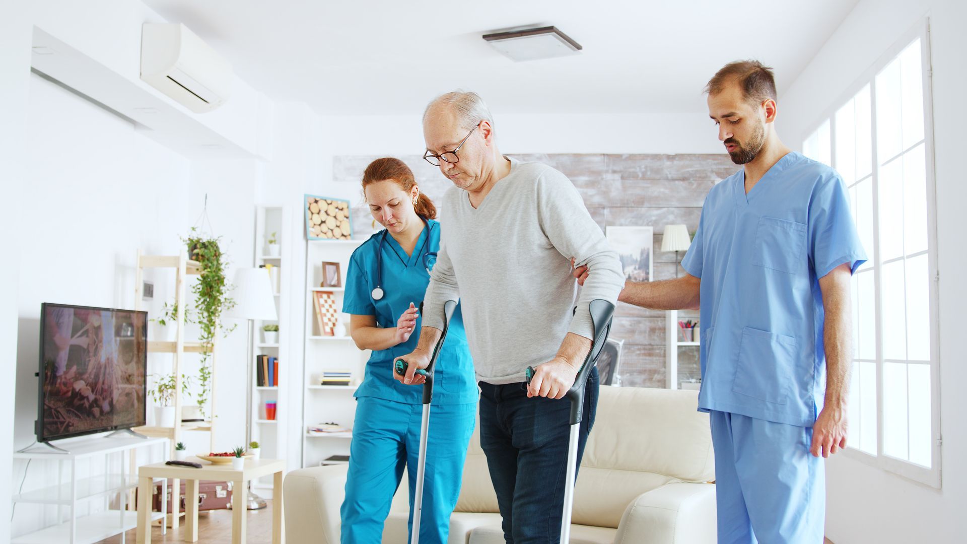 Elderly man with crutches assisted by two medical professionals indoors, walking.