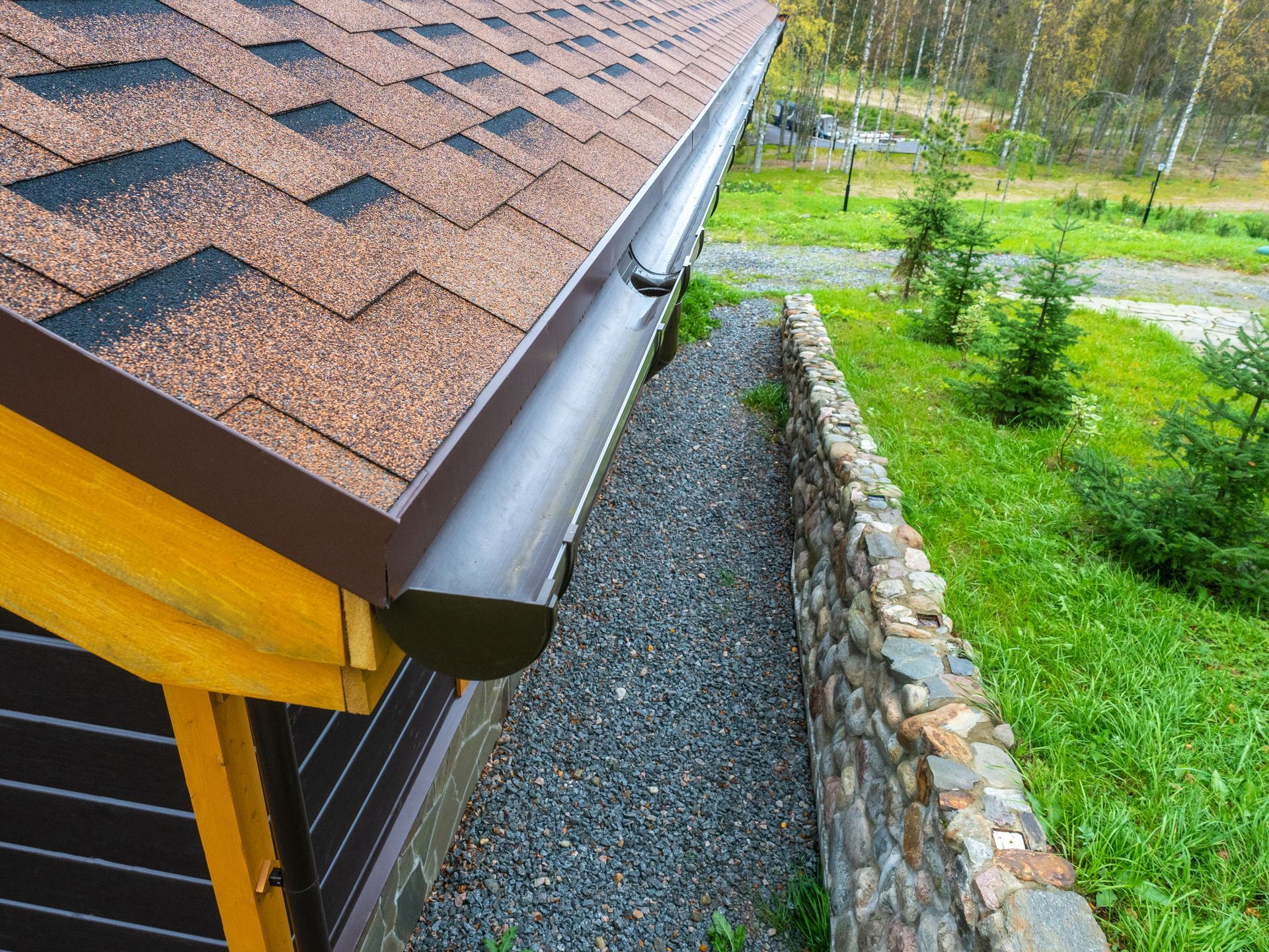 Roof gutter and downspout beside a gravel walkway and stone retaining wall next to a green yard