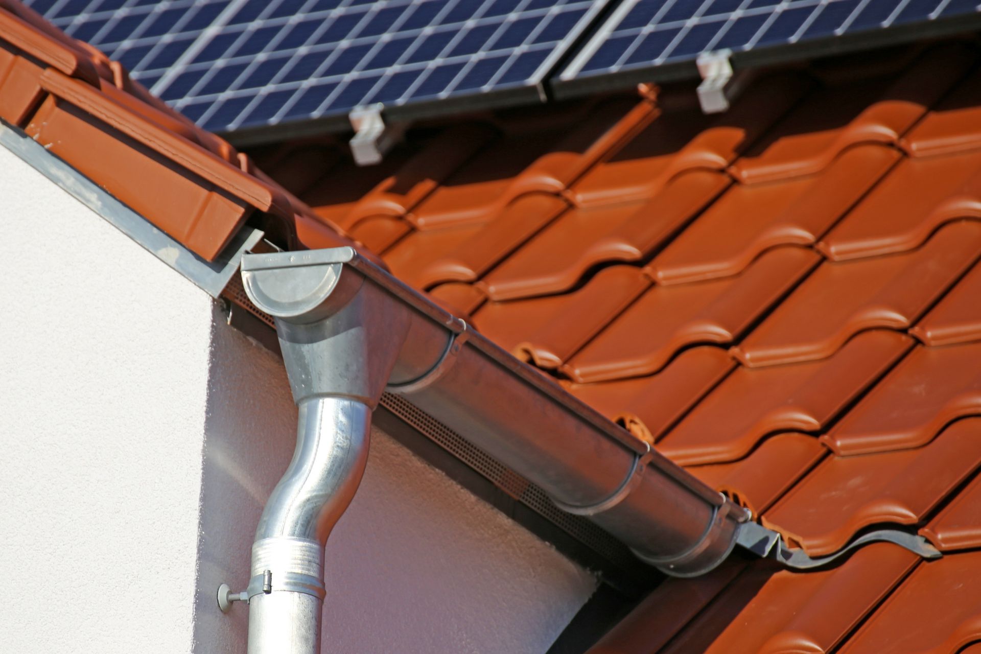 Roof gutter and downspout beside orange tiles, with solar panels above