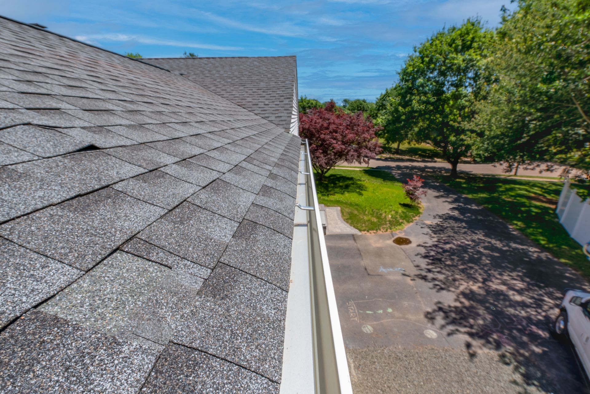 Roof shingles and white gutter beside a driveway, with trees and a blue sky in the background
