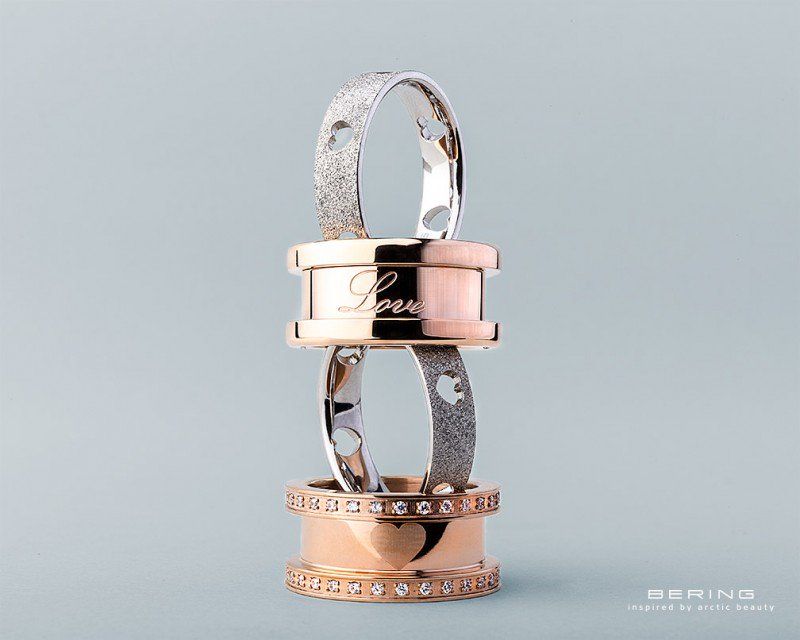 Zwei Bering-Ringsets aus Roségold, eines davon mit einem gravierten „Love“-Band vor einem schlichten hellblauen Hintergrund.