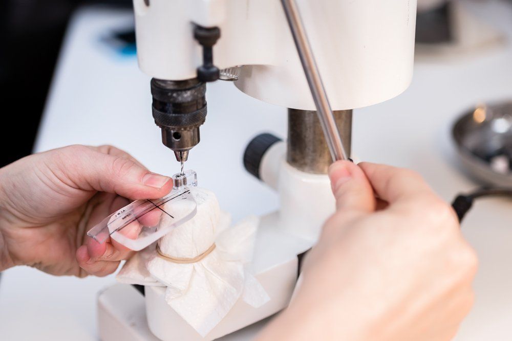 Eine Person bohrt mit einem Präzisionsbohrer ein kleines Loch in ein mit einer Ausrichtungshilfe markiertes Brillenglas.