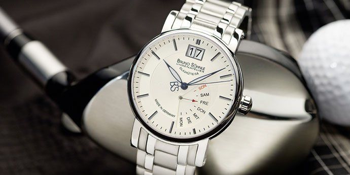 Auf einem Golfschlägerkopf liegt neben einem Golfball eine silberfarbene analoge Armbanduhr mit weißem Zifferblatt.