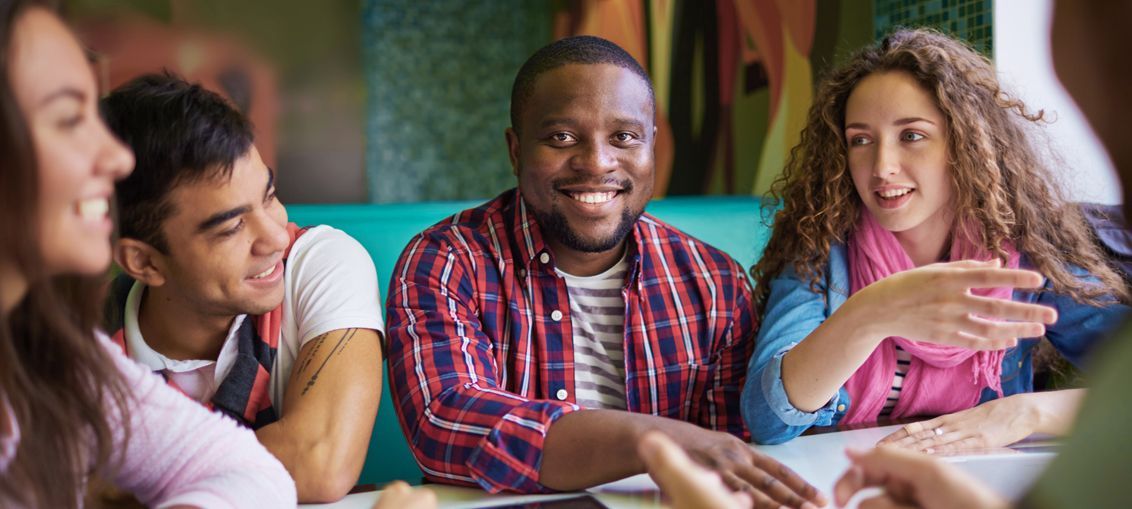 Five people of color smiling together, posing outside with a wooden fence and greenery in the background.