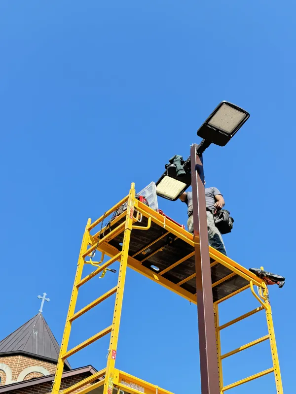 Workers on a yellow scaffolding install outdoor lights on a pole next to a building with a cross, blue sky.