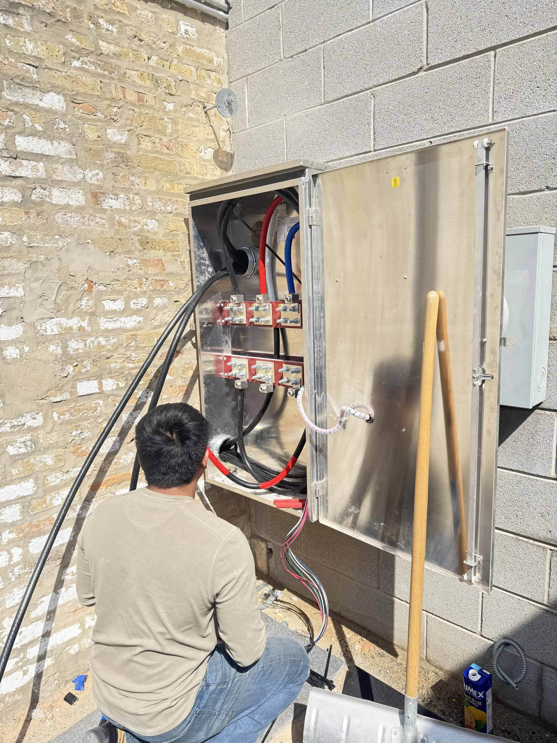 Man working on an electrical box mounted on a brick wall outdoors. Wires, cables, and tools are present.