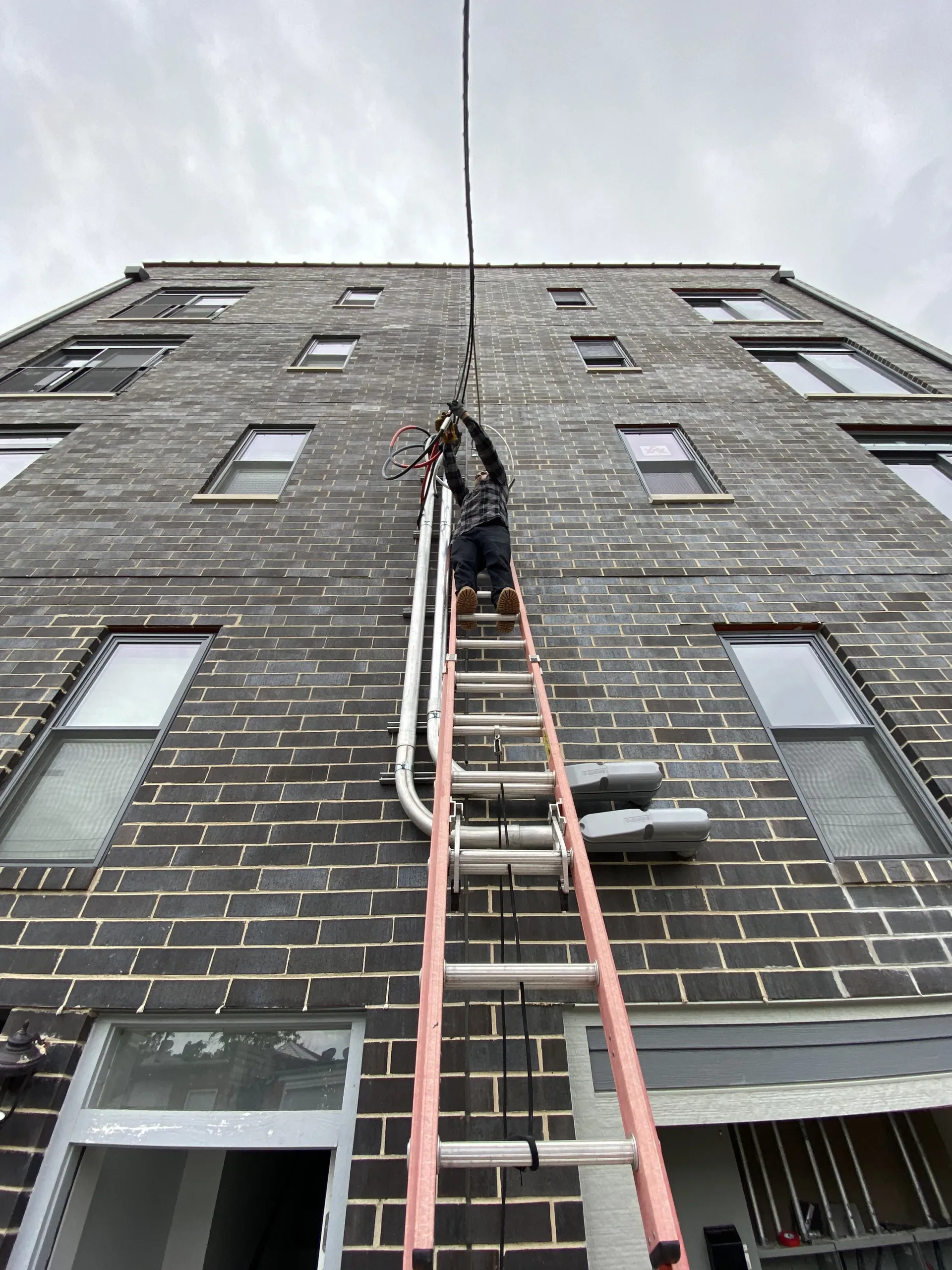 Person on a ladder working on electrical wires attached to a brick building with gray sky.