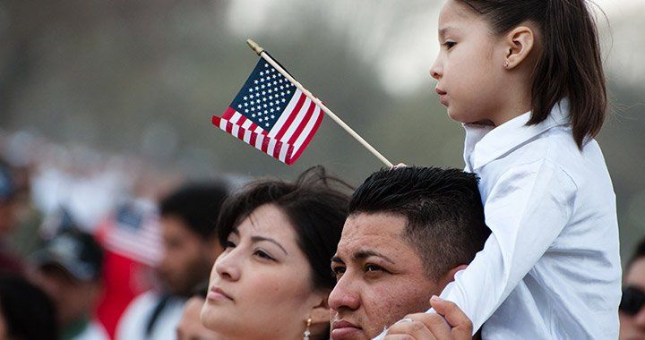 father holds daughter on shoulders while she holds a small american flag