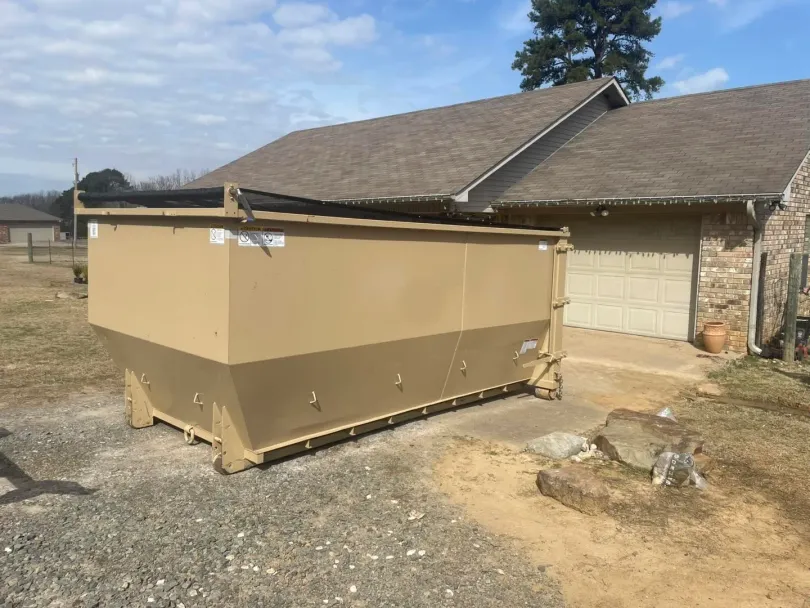 A large, tan dumpster is parked next to a brown-roofed house with a garage. The scene is outdoors on a sunny day.