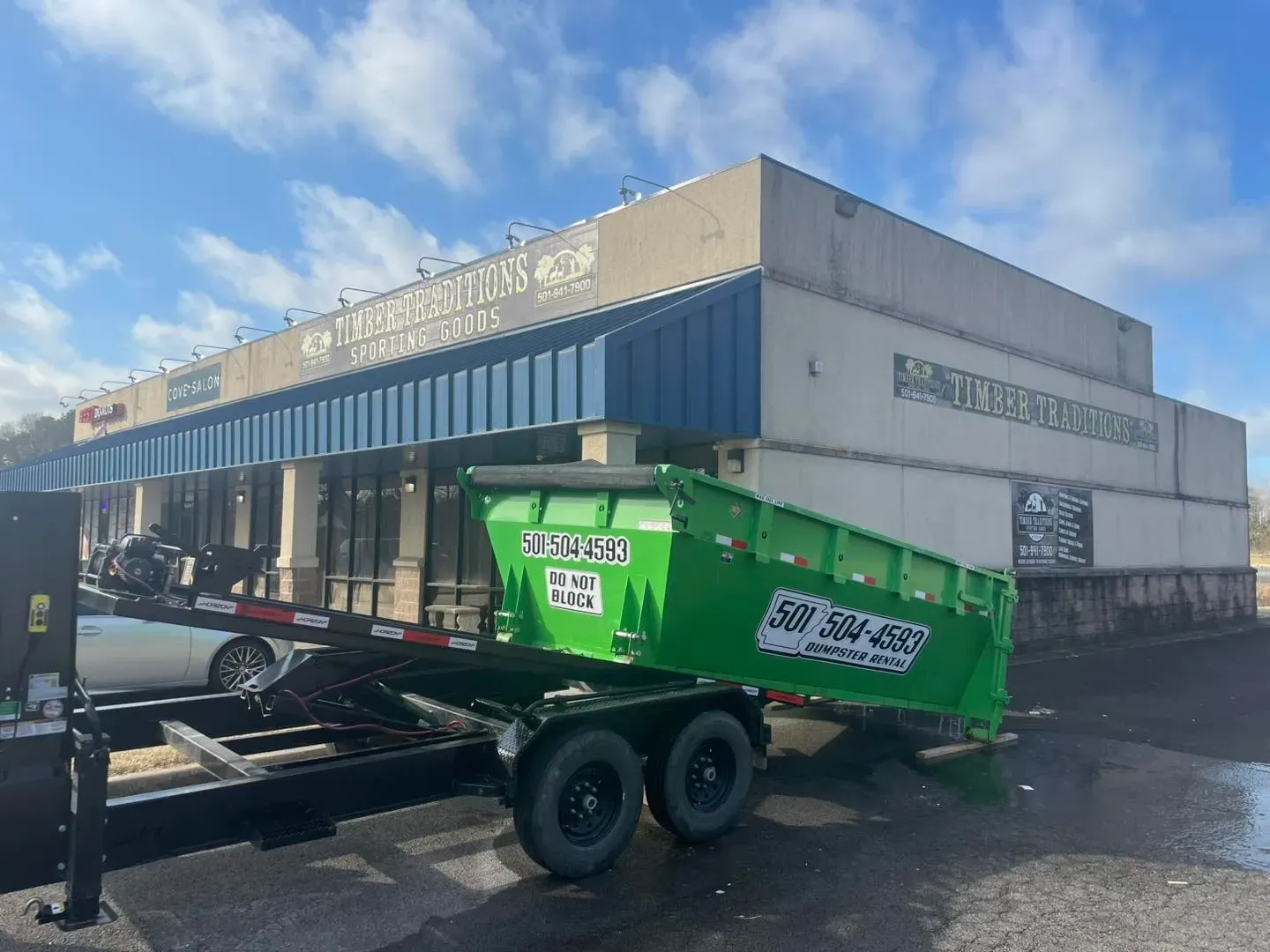 A green dumpster on a trailer in front of a strip mall. The sky is blue and cloudy.