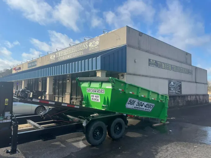 A green dumpster on a trailer in front of a strip mall. The sky is blue and cloudy.