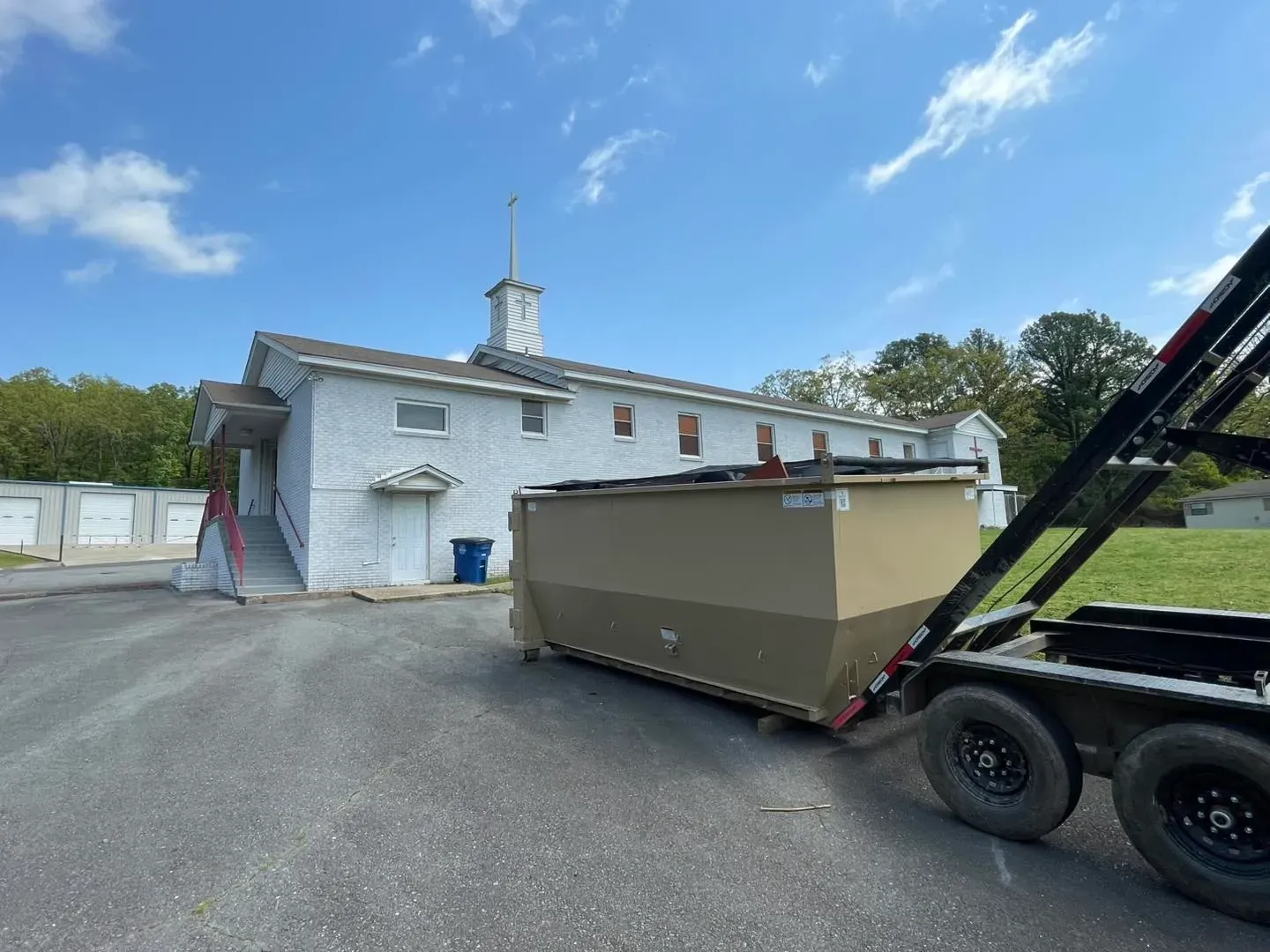 A large dumpster on a trailer sits in front of a white building with a steeple, likely a church, on a sunny day.