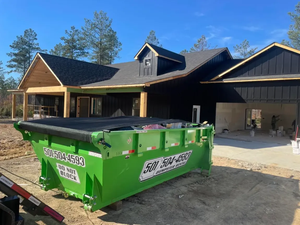Green dumpster in front of a newly constructed black-sided house with wooden trim. The house is on a dirt lot under a blue sky.