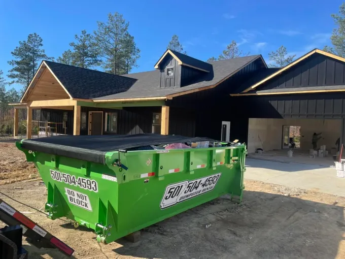 Green dumpster in front of a newly constructed black-sided house with wooden trim. The house is on a dirt lot under a blue sky.