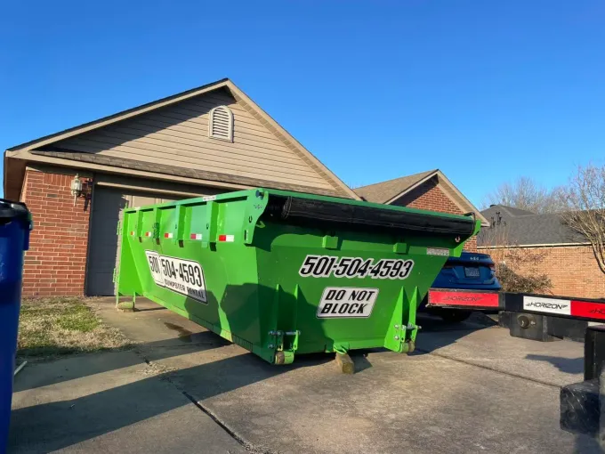 A bright green dumpster parked in a residential driveway. It is in front of a brick house on a sunny day.