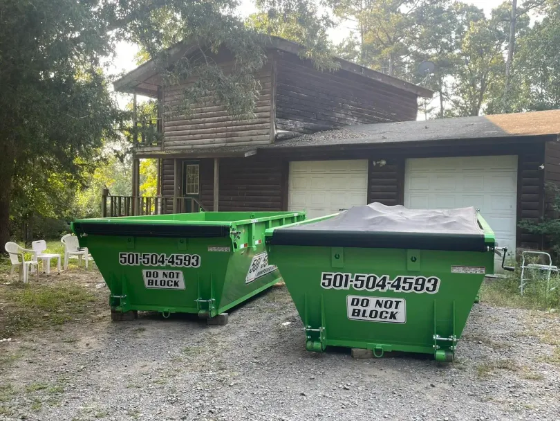 Two green dumpsters in front of a two-story log cabin with two garage doors, warning signs say 