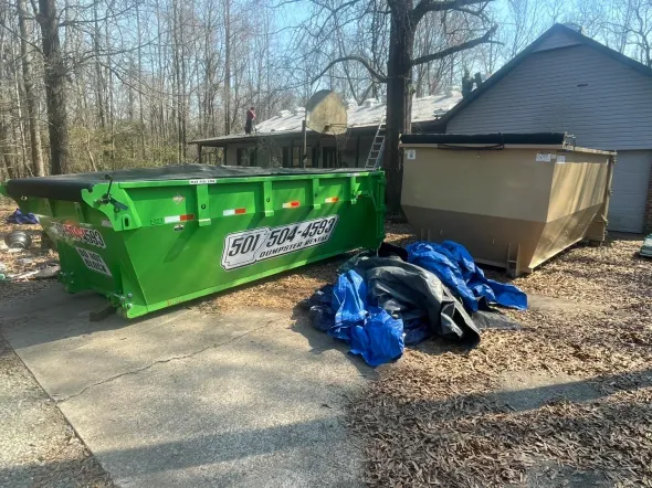 Two dumpsters, one green and one tan, sit on a driveway in front of a house with a damaged roof. Blue tarps lie nearby.