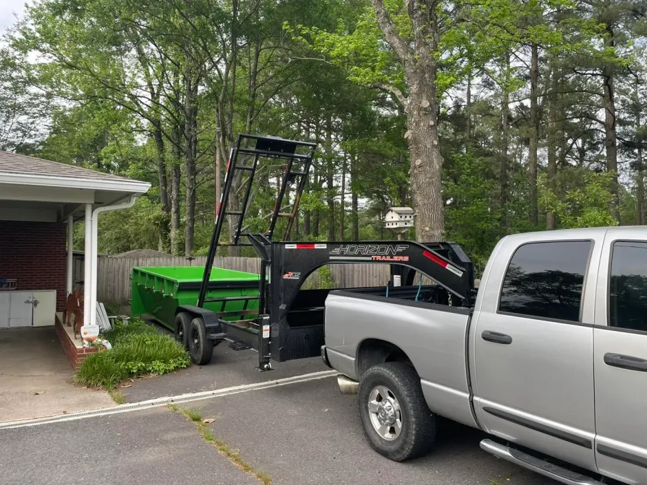 A silver pickup truck is hitched to a large green dump trailer, parked in a driveway.