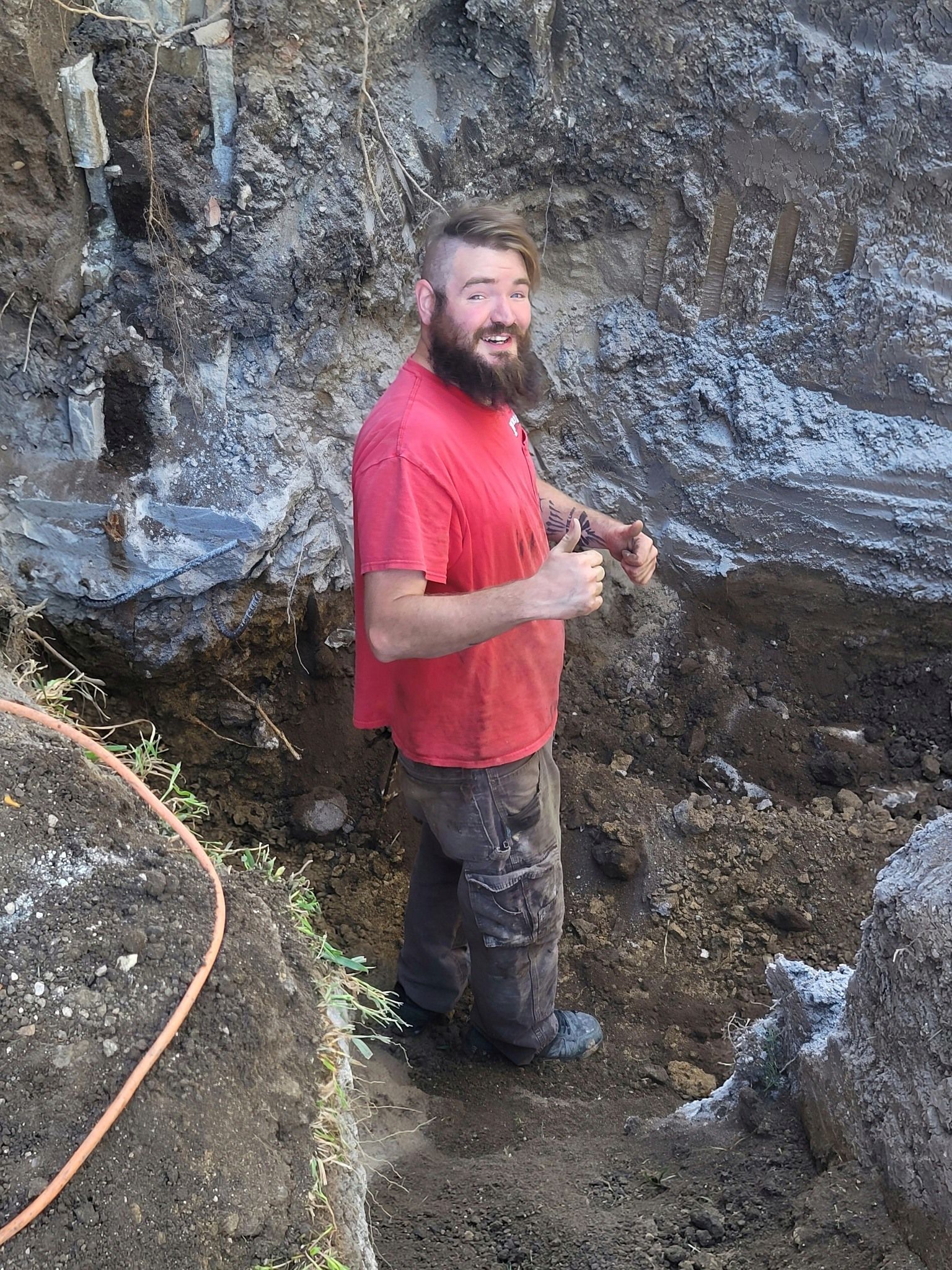 A Man with A Beard Is Standing in The Dirt Holding a Shovel and Giving a Thumbs Up.