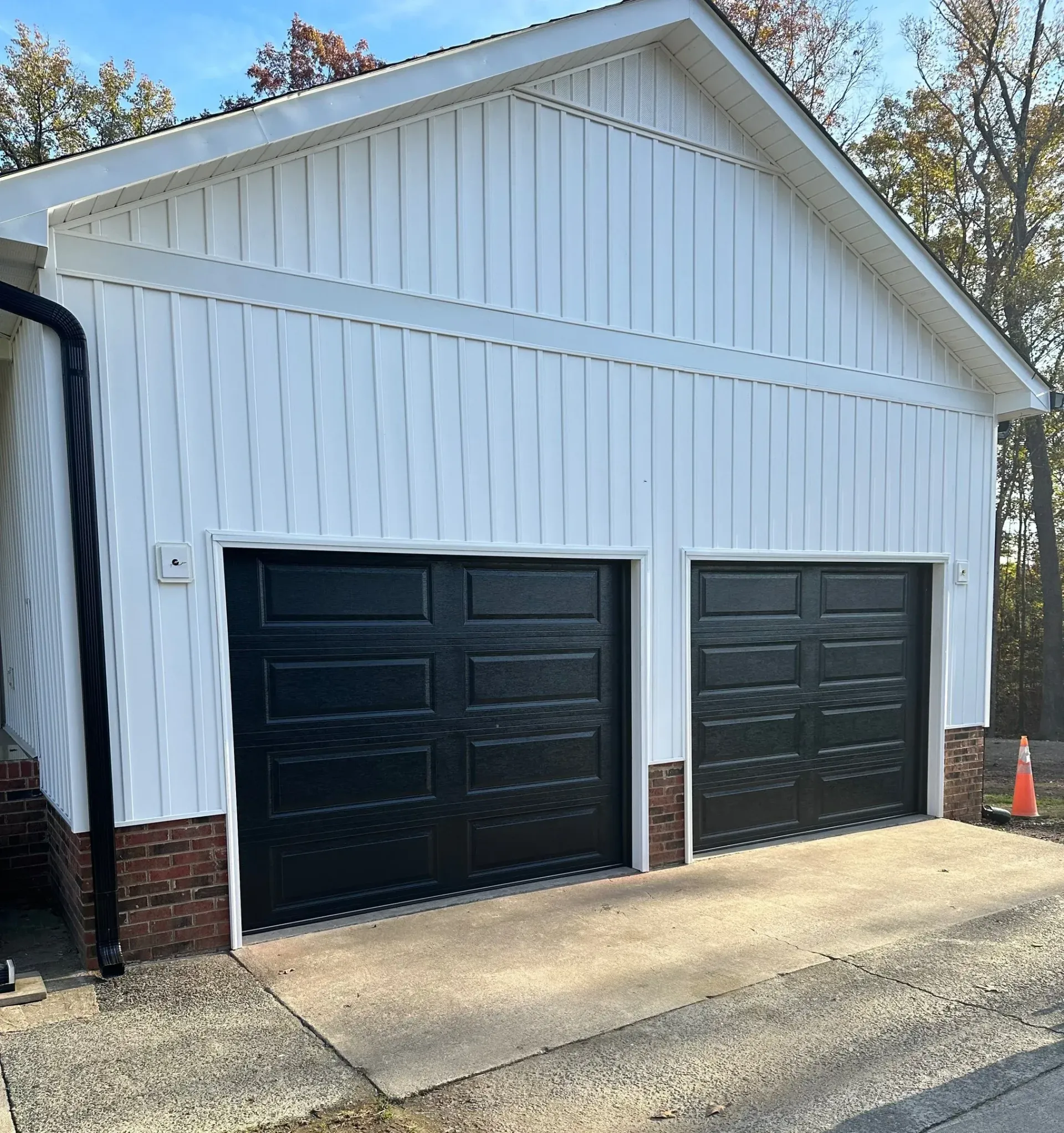 White-sided garage with two black garage doors, brick base, and concrete driveway.