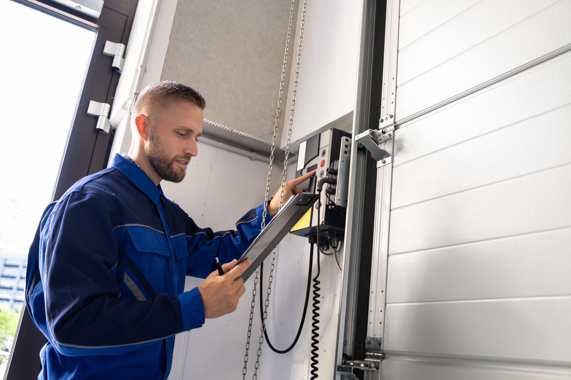 Man in blue overalls inspects a garage door control panel, using a clipboard.