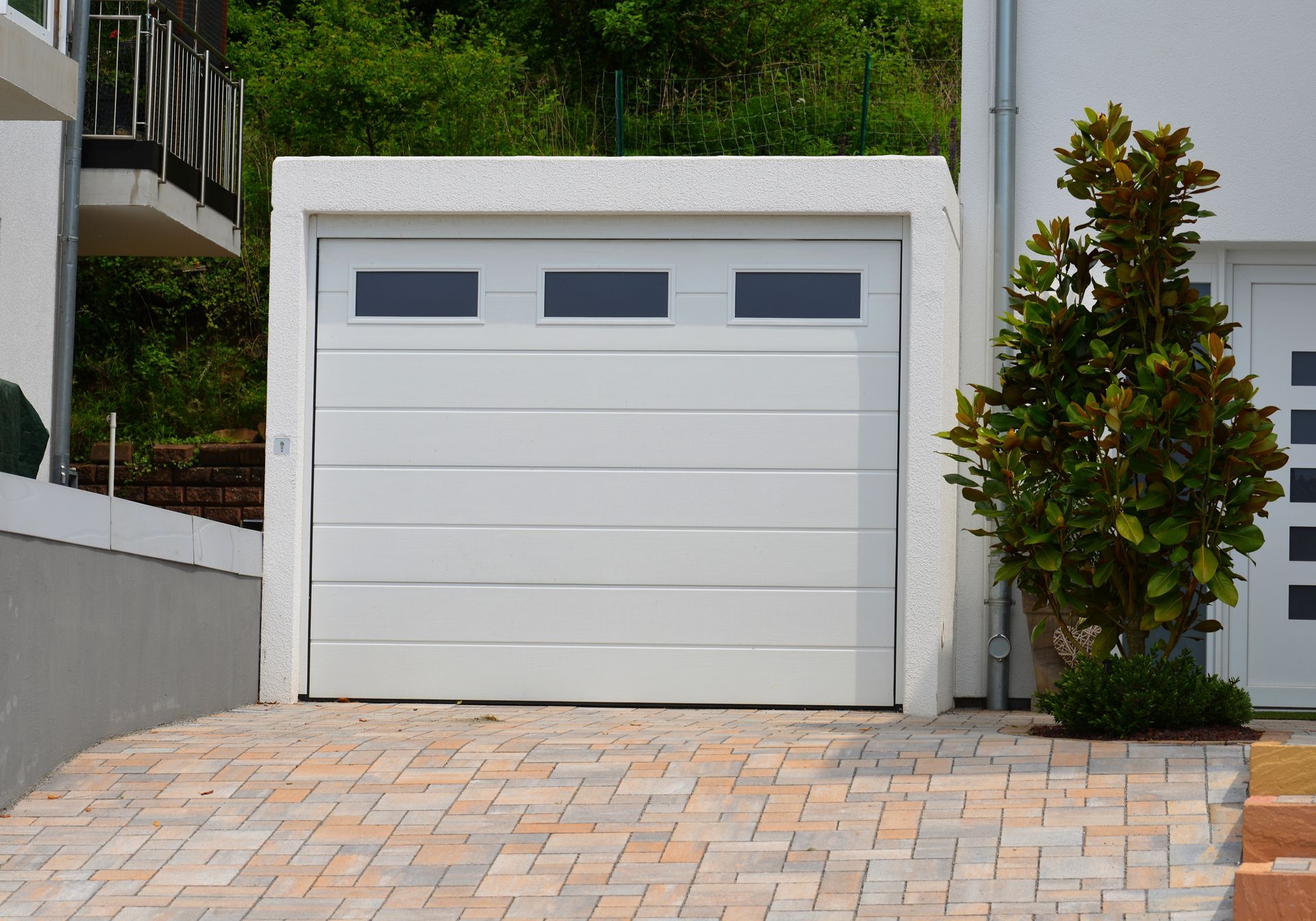 White garage door with three upper windows, set in a stone-framed opening; brick driveway, small tree.