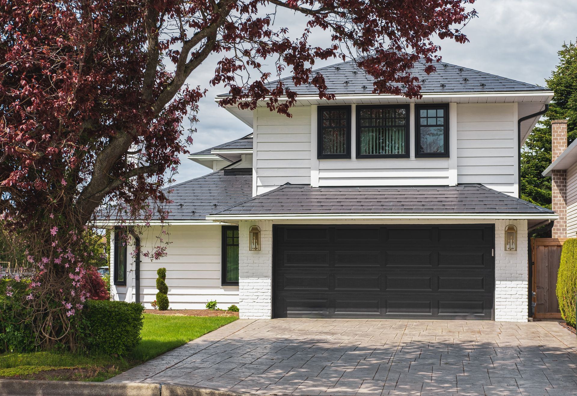White house with black garage door, driveway, and dark roof; a red-leafed tree is in the foreground.