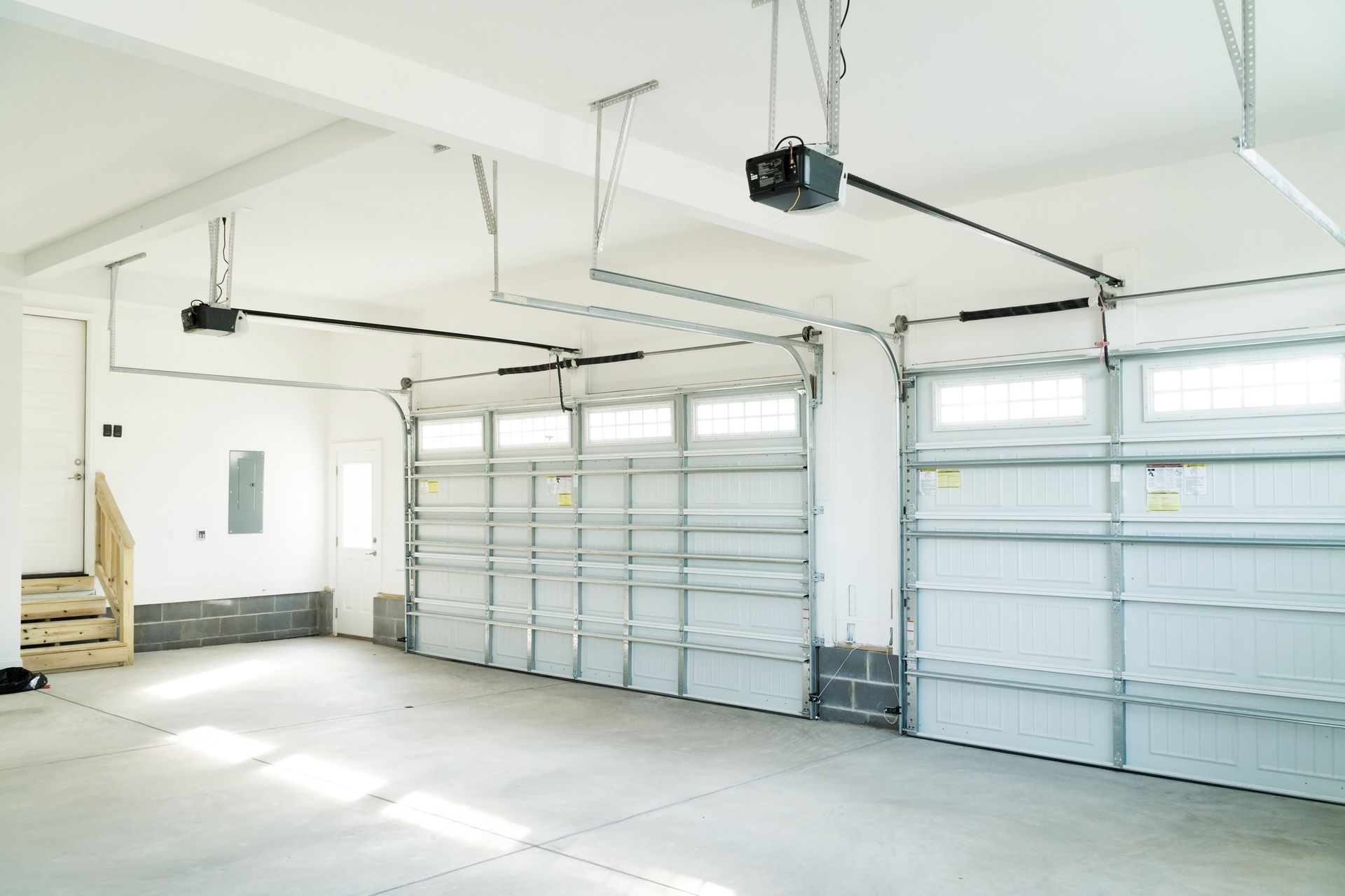 Bright, empty garage with two closed doors, ceiling-mounted openers. Concrete floor, white walls.