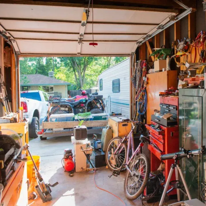 Cluttered garage with open door; trailer with ATV, camper, bicycle, tools, and a truck parked outside.
