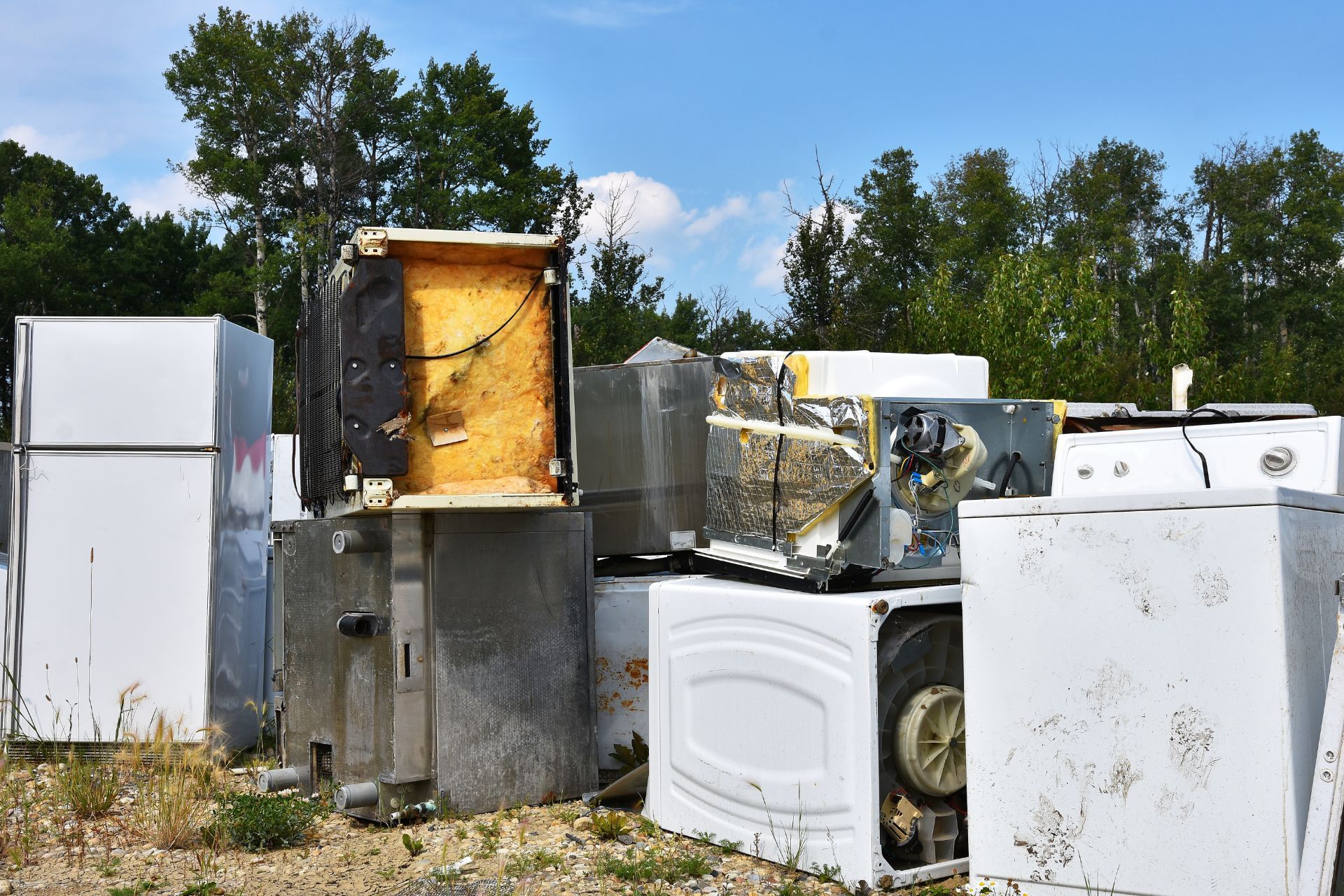Discarded appliances, including refrigerators and washing machines, piled outdoors in a junkyard under a blue sky.