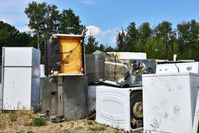 Discarded appliances, including refrigerators and washing machines, piled outdoors in a junkyard under a blue sky.