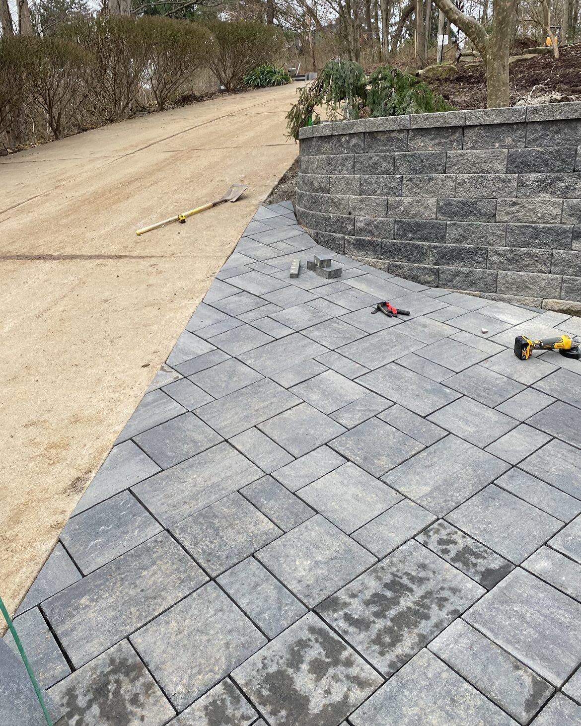 Asphalt paving with gray stone bricks curving toward a retaining wall. A dirt road and bushes are in the background.
