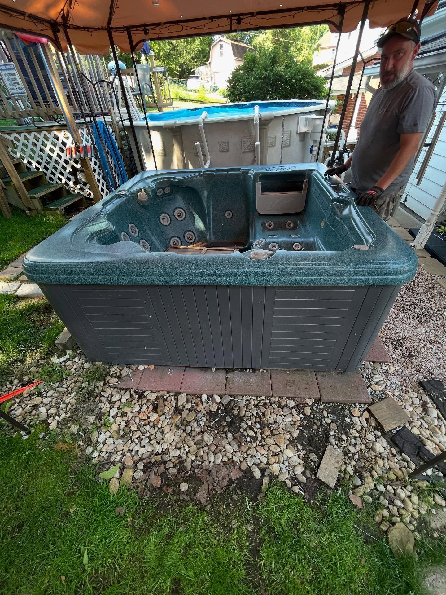 A person standing near a hot tub on a brick-lined surface outdoors, under a canopy. The hot tub is dark green.