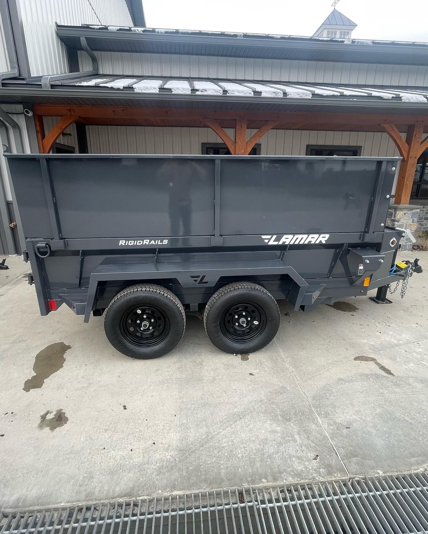 Dark gray Lamar dump trailer with black wheels, parked on a concrete surface, in front of a wooden-trimmed building.