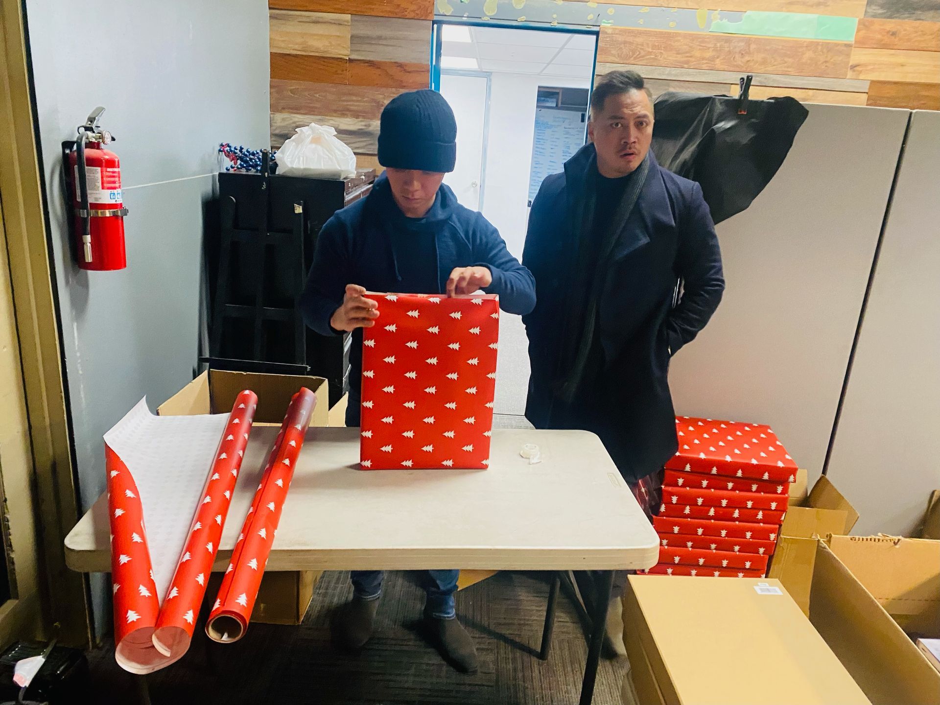 Two men are standing at a table wrapping gifts.