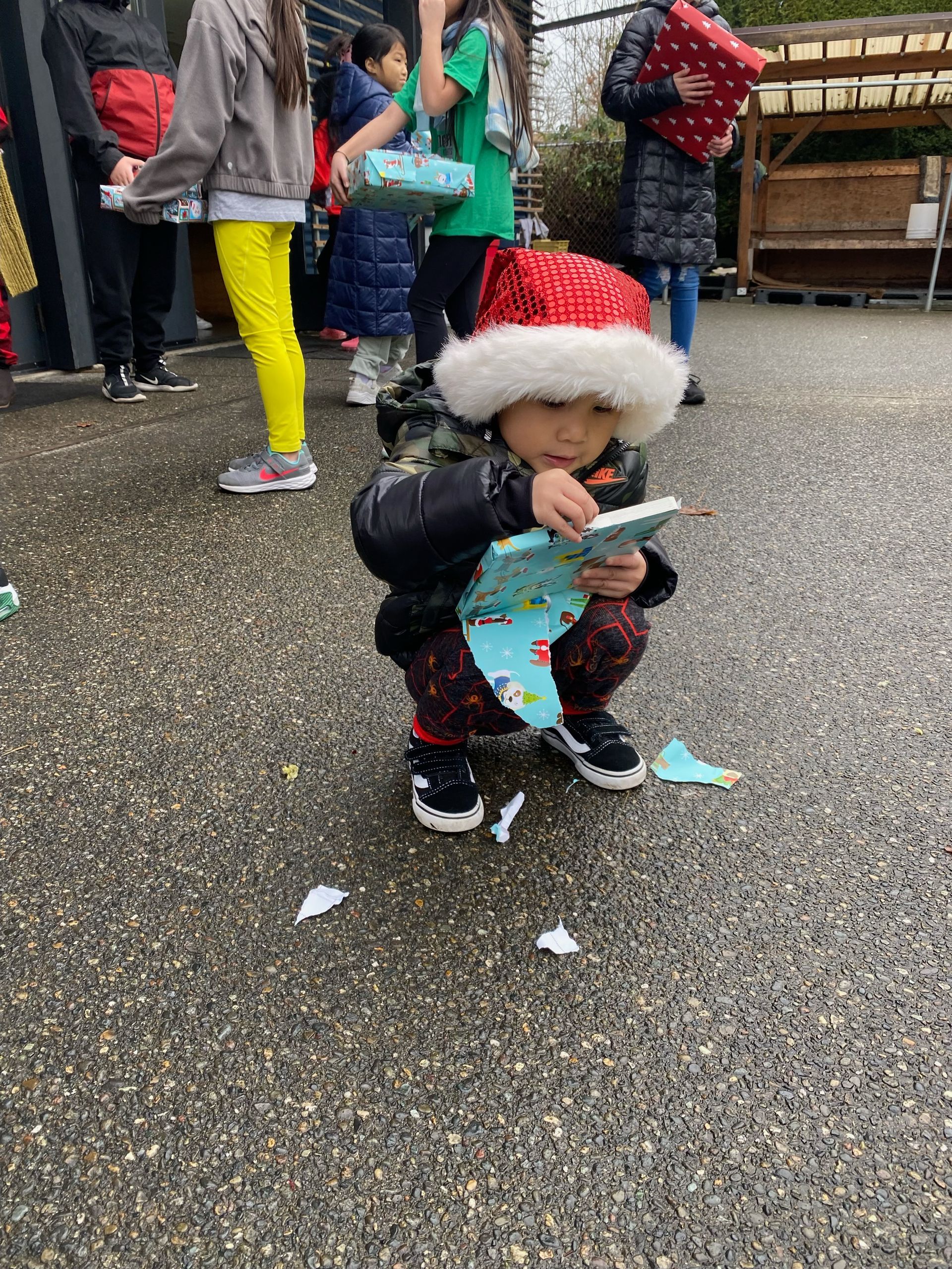 A little boy wearing a santa hat is kneeling down on the ground holding a piece of paper.