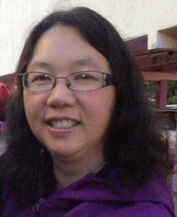 A woman wearing glasses and a purple jacket is smiling.