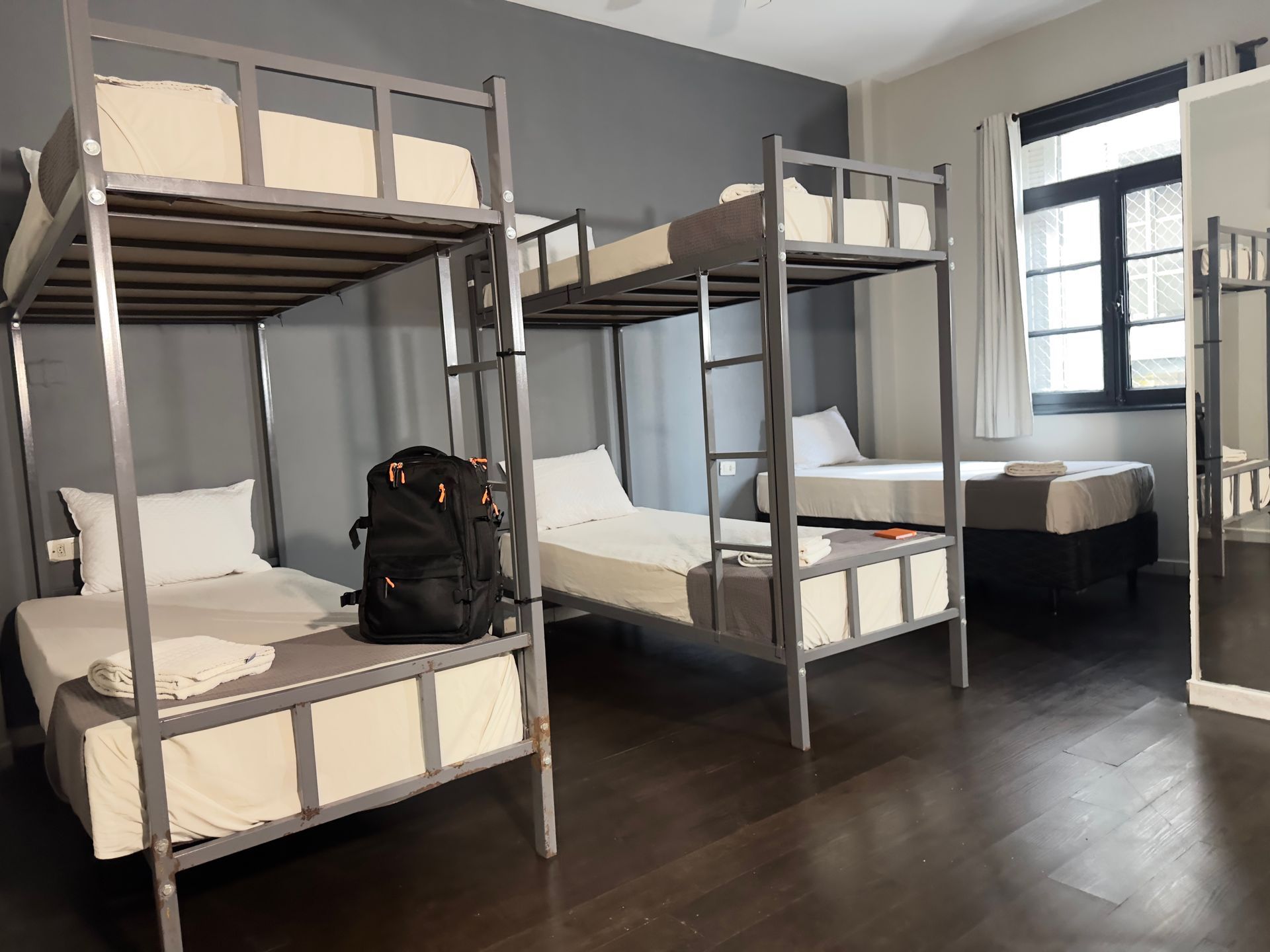 Dormitório de albergue com beliches, mochila e janela. Esquema de cores cinza e branco.