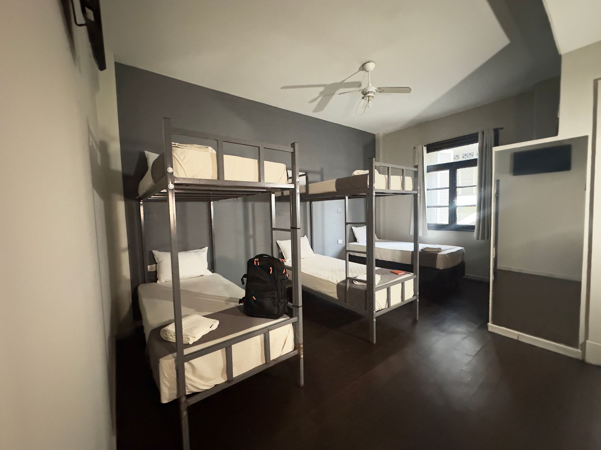 Um dormitório com três beliches, uma janela e um espelho. As paredes são cinza e brancas.