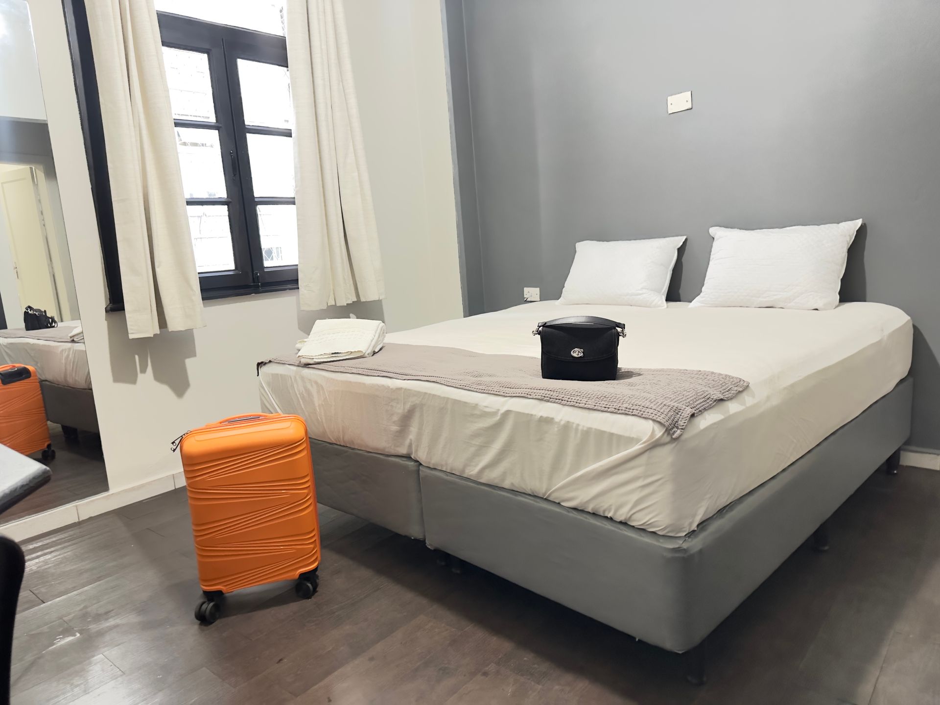 Quarto com cama de casal, mala laranja e janela. Decoração em cinza e branco.