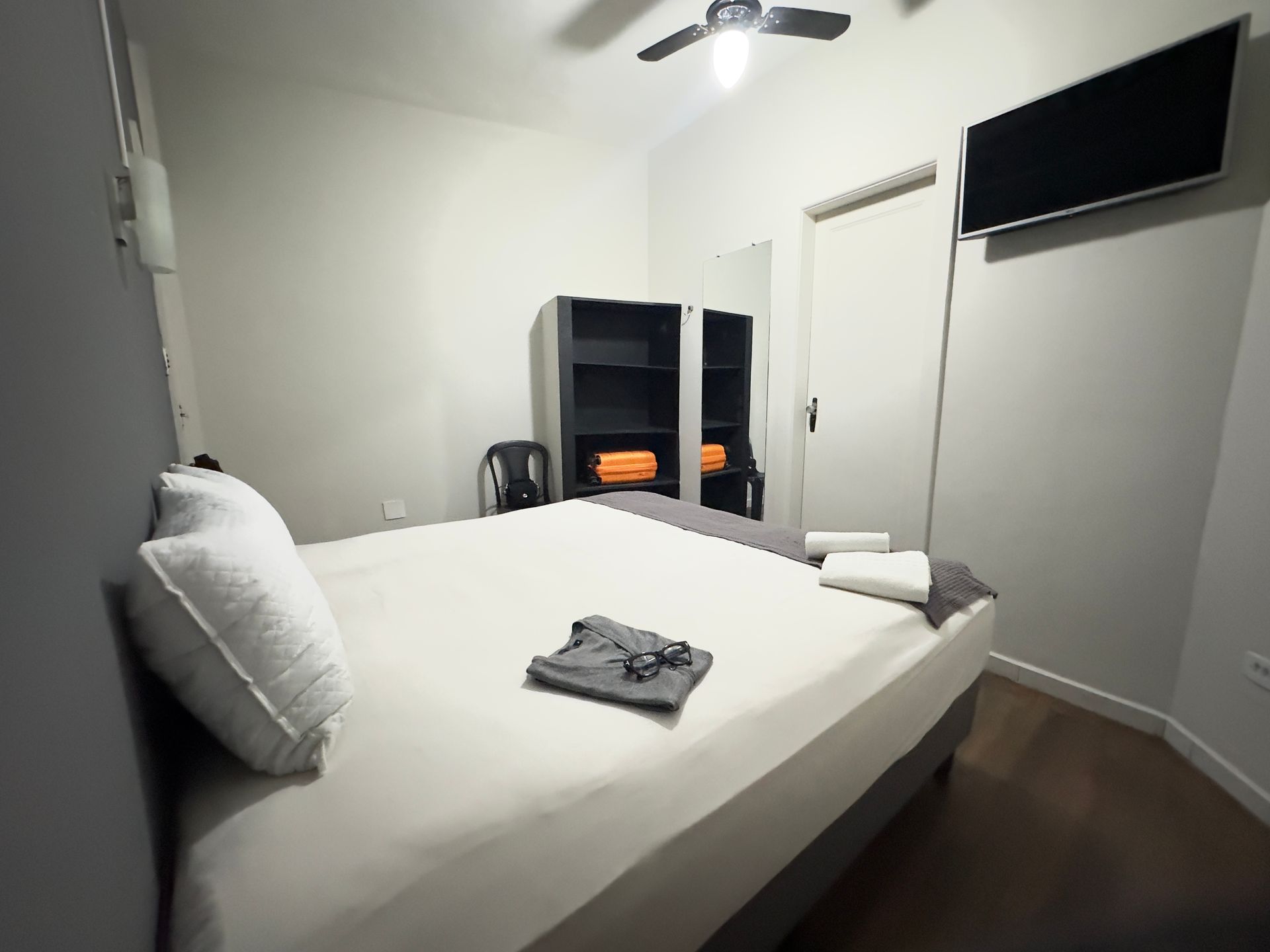 Quarto com cama, TV, armário e toalhas. Combinação de cores cinza e branco.