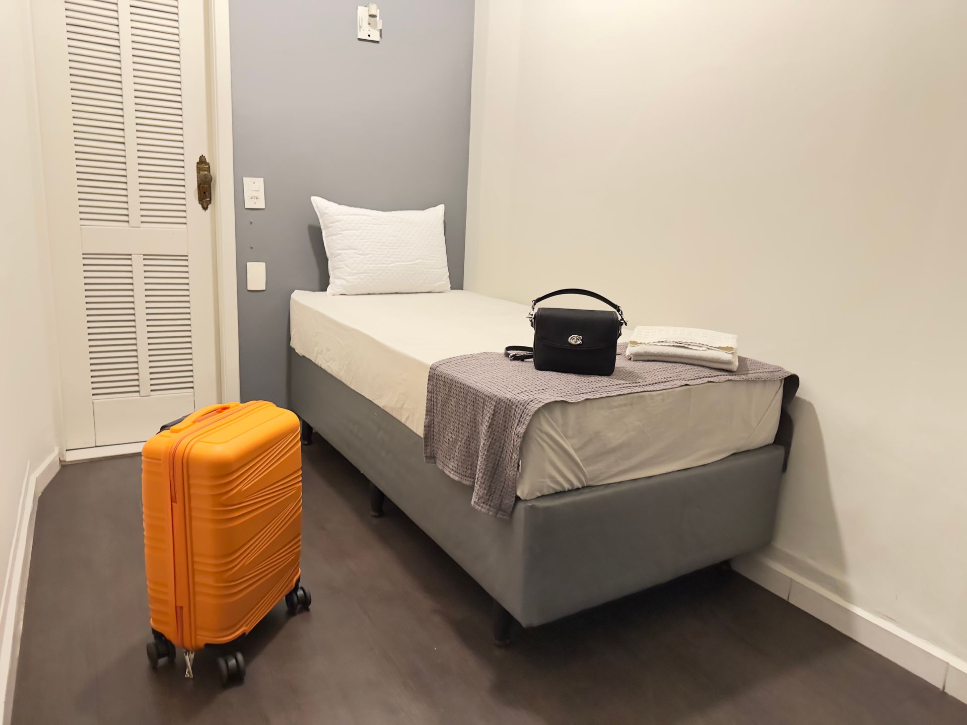 Pequeno quarto de hotel com cama de solteiro, mala laranja e câmera na cama.