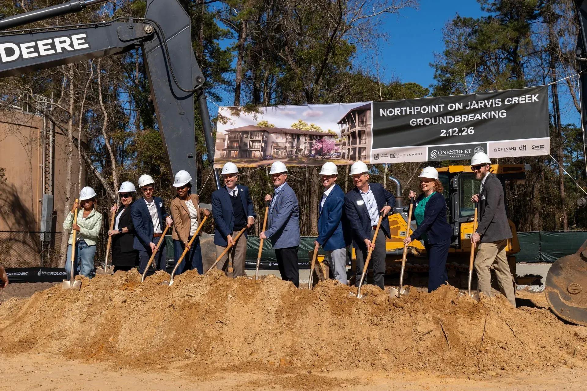 People with hard hats and shovels at a groundbreaking ceremony beside a DEERE excavator and signboard.