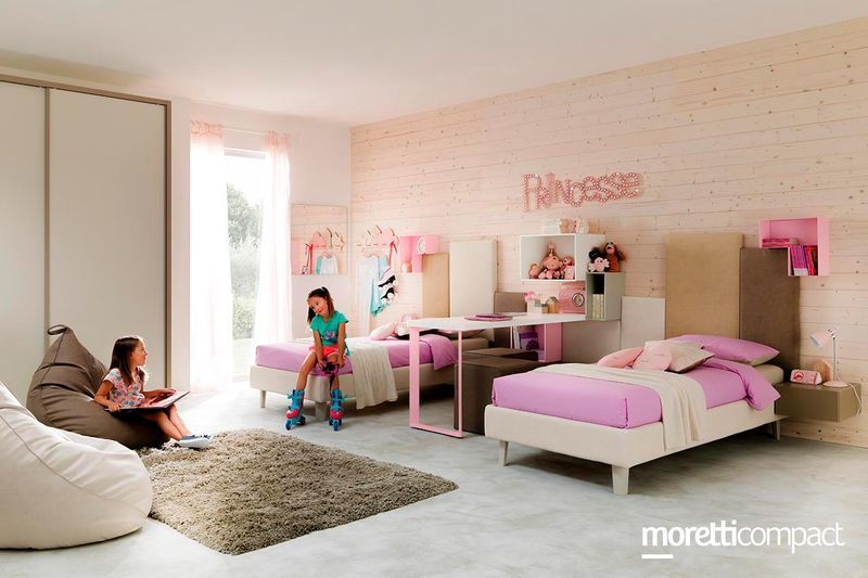 Camera da letto rosa con letto, scrivania e due persone sedute sul pavimento e su una sedia vicino a una finestra.
