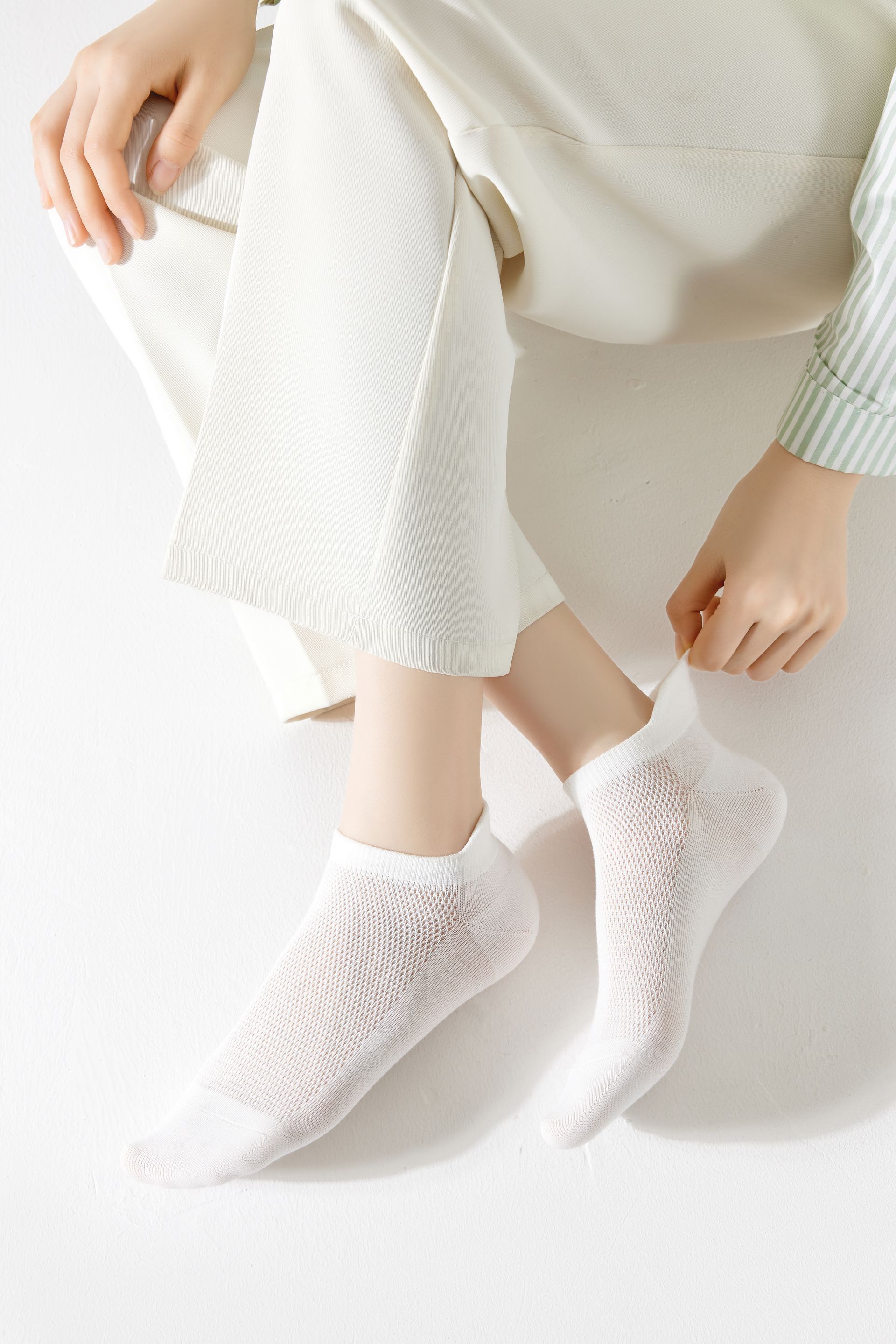 White ankle socks on a person, seated and adjusting the sock. White pants and background.