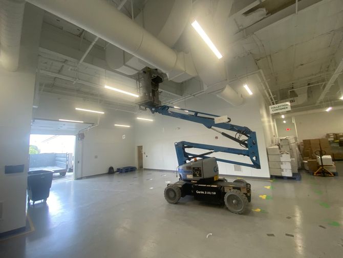 Blue boom lift inside a white warehouse, reaching up to a person working near the ceiling.