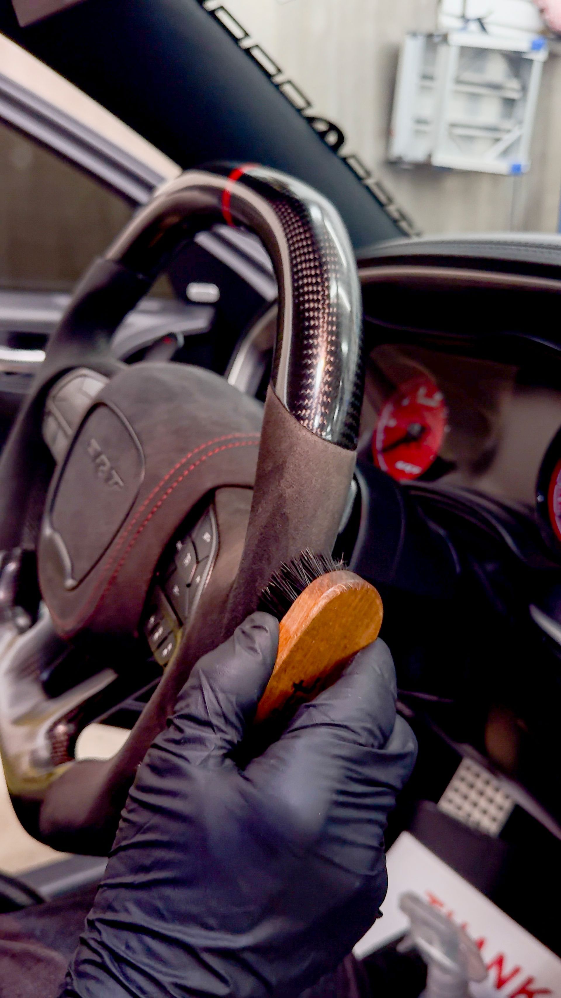 Gloved hand brushes a car's suede steering wheel with a wooden brush; red dashboard and carbon fiber accents visible.