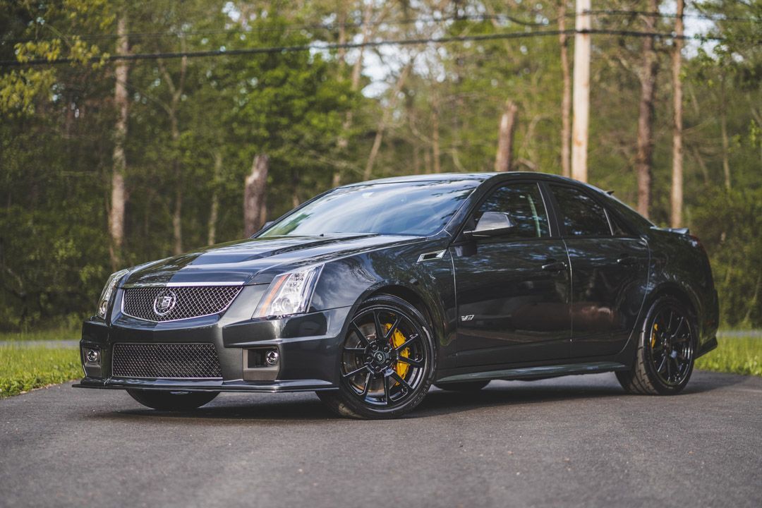 Black Cadillac CTS-V on a paved road, yellow brake calipers, in front of trees.
