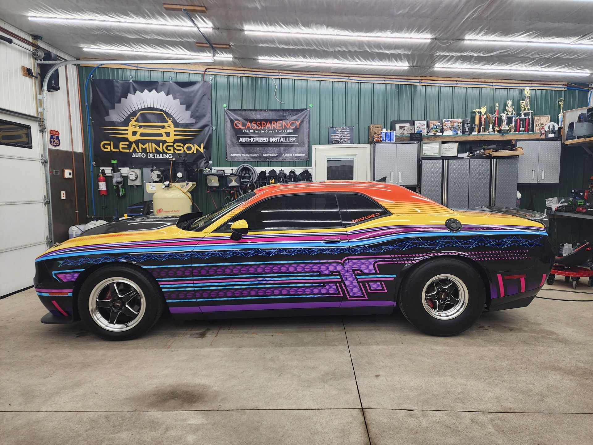 A colorful drag racing car inside a garage. The car has a rainbow wrap and large rear tires.